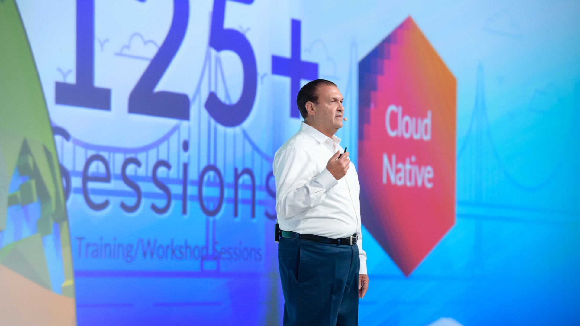 A photo of the event presentation design for DevOps World 2018. It depicts a presenter standing on stage with a wide screen presentation slide behind him.