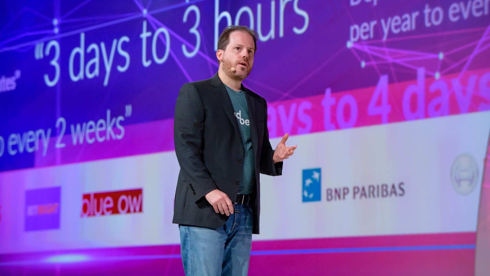 A photo of the event presentation design for DevOps World 2018. It depicts a presenter standing on stage with a wide screen presentation slide behind him.