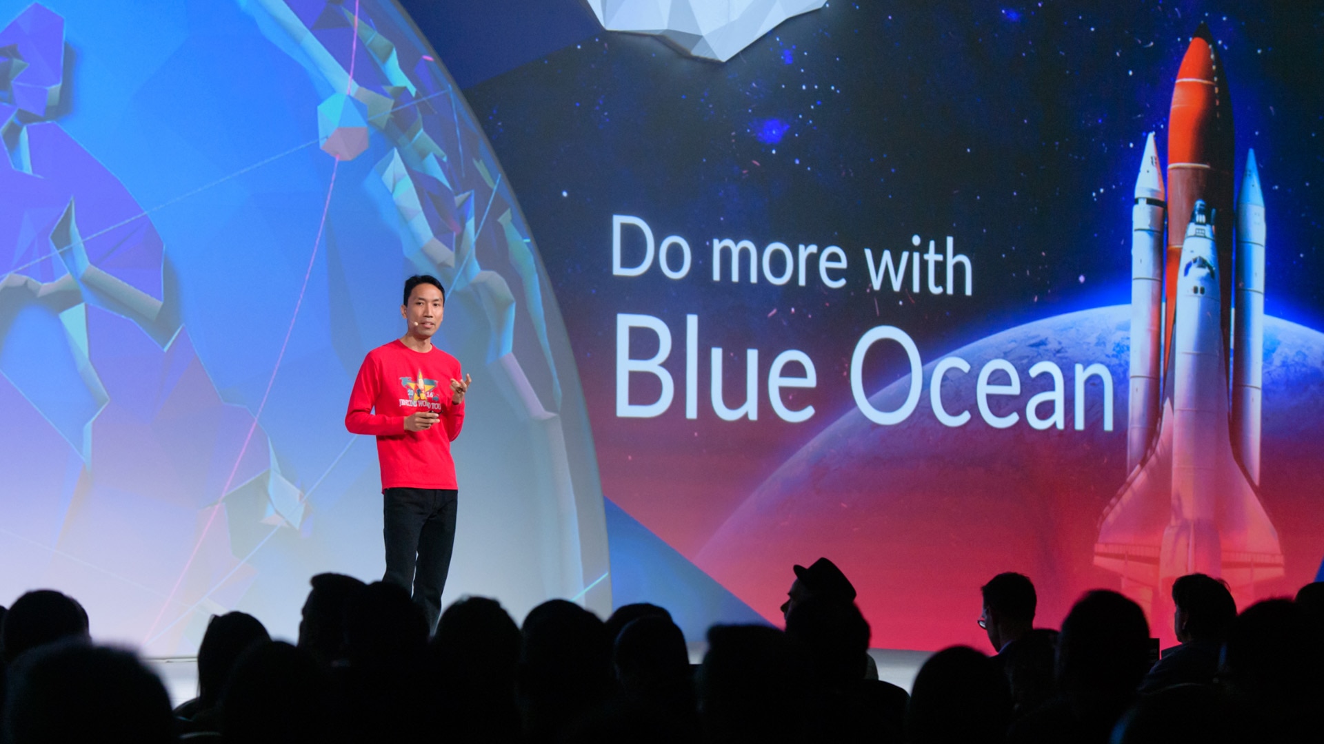 A photo of the event presentation design for DevOps World 2018. It depicts a presenter standing on stage with a wide screen presentation slide behind him.